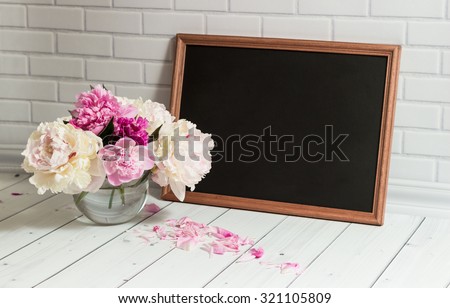 Beautiful bouquet of pink and white peonies in the glass vase with petals and black chalkboard with empty place for your text on the light grey brick and wood background.