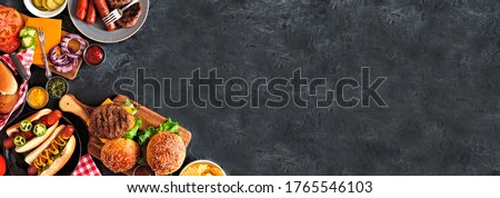 Summer BBQ food corner border with hot dog and hamburger buffet. Top down view over a dark slate banner background. Copy space.
