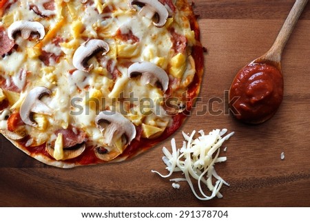 Pizza with ham, mushroom and pineapple against a wood background with shredded mozzarella cheese and spoon of tomato sauce