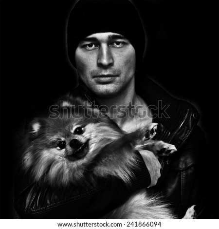 Brutal man in a hat and leather jacket with a dog in her arms on a black background