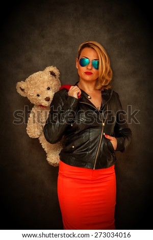 Glamour beautiful girl-blonde in a leather jacket, sunglasses and a red skirt with a teddy bear on a dark background