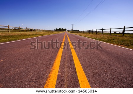 A long road bordered by farm fences and telephone wire against a blue sky