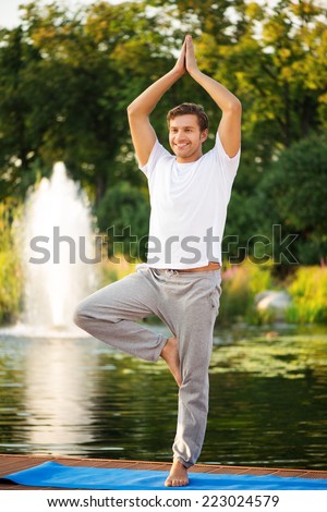 Smiling young man practicing yoga, doing tree pose. With a beautiful green scenery of park on background