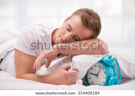 Close up photo of tired and sleepy young man. He lying in white bed with blue alarm clock under pillow