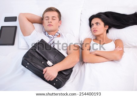 Top view photo of young couple lying in bed under white blanket with electronic gadgets. Angry woman looking at sleeping husband who holding briefcase