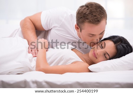 Young couple lying in white bed early in the morning. Handsome man kissing his sleeping wife