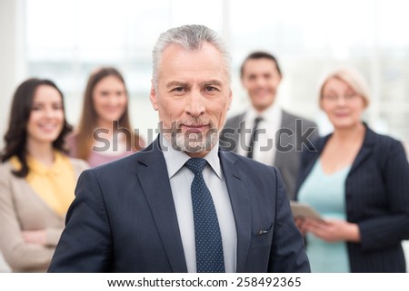 Businessman looking at camera. His team standing behind. Office interior with big window