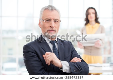 Businessman looking at camera. His assistant holding a stack of papers. Office interior with big window