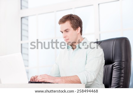 Young businessman working with laptop and sitting at his black leather office chair. Office interior with window