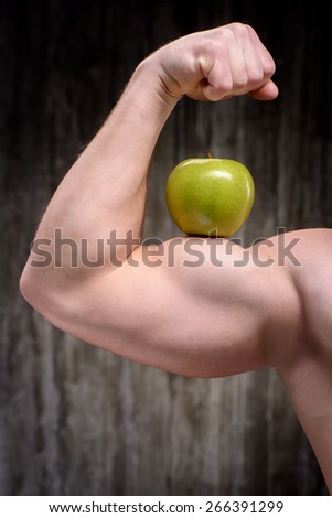 Close up photo of young well formed sporty man demonstrating biceps with green apple on it. Fitness and healthcare concept