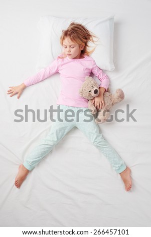Top view photo of little girl sleeping on white bed with teddy bear. Quiet Freefaller pose. Concept of sleeping poses