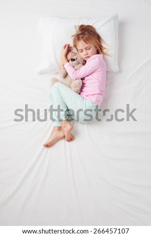 Top view photo of little girl sleeping on white bed with teddy bear. Quiet Foetus pose. Concept of sleeping poses