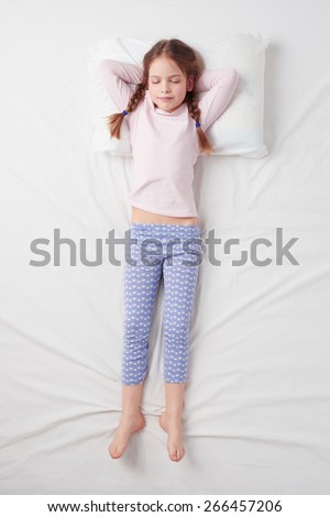 Top view photo of little cute girl sleeping on white bed with arms over the head. Quiet Soldier pose. Concept of sleeping poses