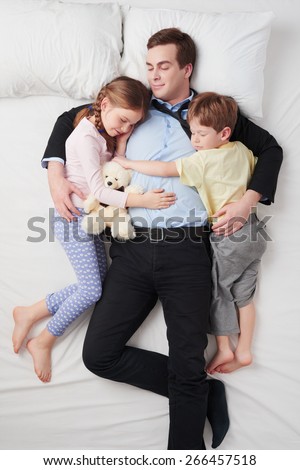 Top view photo of tired businessman wearing suit, and his two children. Father\'s arms are over daughter and son. They sleeping on white bed and hugging each other
