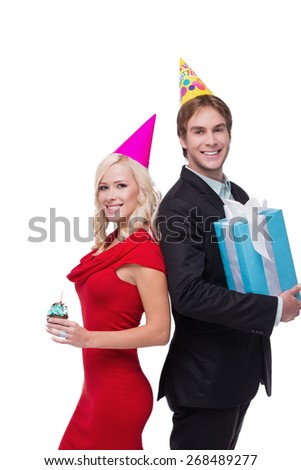 Photo of young smiling man and woman looking at camera with cupcake and present. They wearing birthday hat and standing on white background. Concept for happy birthday