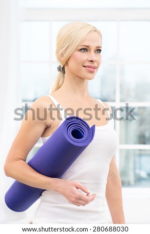 Close up photo of beautiful sporty woman. Woman holding violet fitness mat and smiling. White interior with large window
