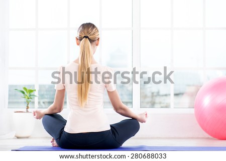 Nice photo of young woman practicing yoga. Woman meditating while doing lotus pose and turning back to camera. White interior with large window