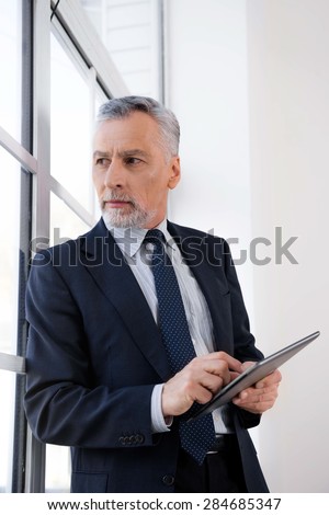 Businessman in years standing near large window, holding tablet computer and staring at window