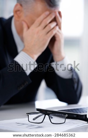 Disappointed businessman in years sitting near notebook and glasses. Man covering his face with hands