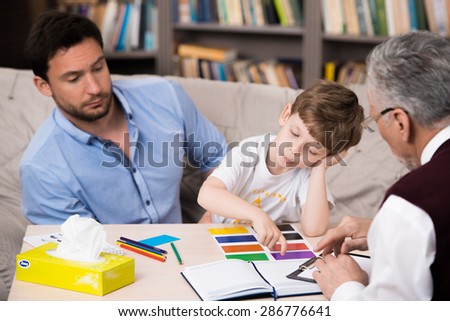Little boy and his father talking with psychologist. Psychologist making test with boy. Sad boy choosing color cards. There are many books in psychologist office