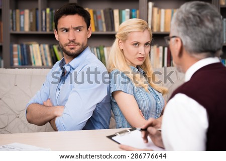 Young couple talking with psychologist. Psychologist taking notes. Angry man and woman sitting back to back. There are many books in psychologist office