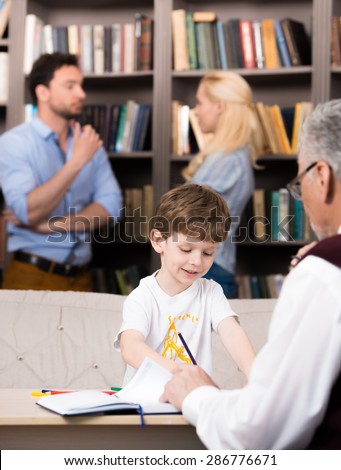 Young family of three talking with psychologist. Psychologist taking notes. Boy trying to draw something. Parents arguing. There are many books in psychologist office