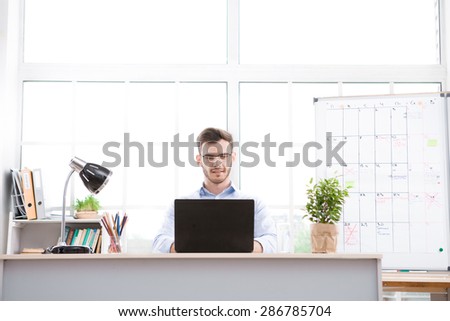 Young businessman wearing glasses is in office full of folders for documents and office supplies. Businessman using laptop
