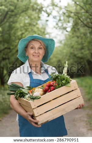 Good-looking smiling middle-aged woman-farmer holding wooden box with fresh vegetables and standing in green garden