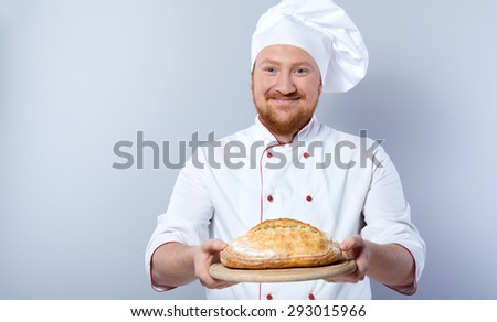 Portrait of positive young male chef in white uniform. Head-cook smiling, looking at camera and proposing freshly baked bread. Standing against grey background
