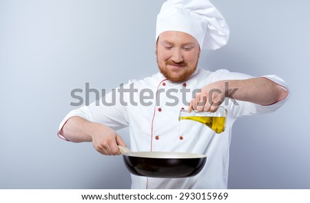 Portrait of positive young male chef in white uniform. Head-cook holding pan and pouring olive oil in it. Standing against grey background