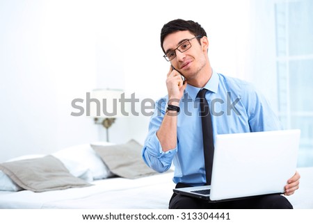 Young smiling businessman using phone and laptop while sitting in cozy hotel room