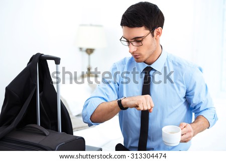 Young businessman finishing his coffee and looking at watch while sitting in cozy hotel room with suitcase