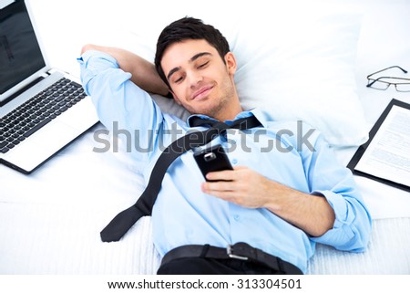 Young smiling businessman lying in cozy hotel room on big white bed with laptop, documents and using phone