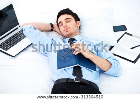 Young businessman napping in cozy hotel room on big white bed after reading book. Laptop and phone are near him