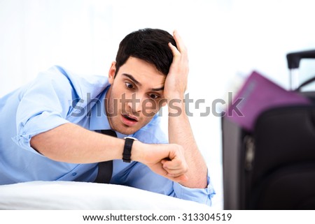 Young shocked businessman looking at watch while being late. He is in cozy hotel room with suitcase