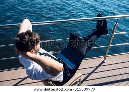 Photo of handsome young businessman sitting and relaxing near river. Man wearing suit and using laptop