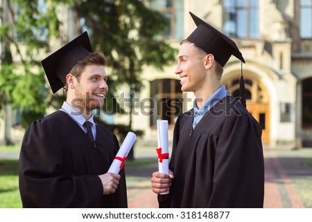 Young male students dressed in black graduation gown. Campus as a background. Boys cheerfully smiling, holding diplomas and looking at each other