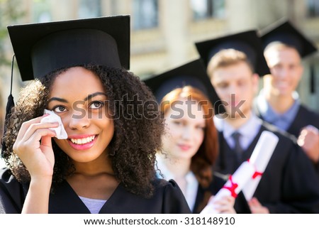 Young students dressed in black graduation gown. Campus as a background. Afro American girl crying, smiling, holding diploma and looking aside. Students are on background standing in row