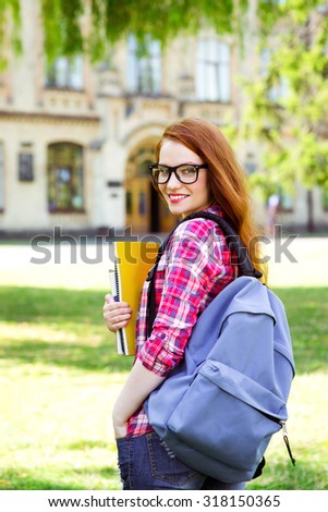Photo of young female student with backpack, glasses and books. Campus as a background. Girl smiling and looking at camera