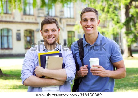 Photo of young male students with backpack and notebooks. Campus as a background. Boys with cup of coffee smiling and looking at camera
