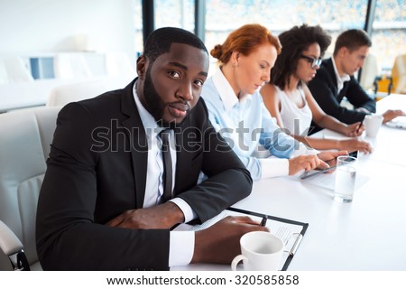 Photo of creative multi ethnic business group. Mixed race business team using electronic devices and working. Accent on African businessman. White modern office interior with big window