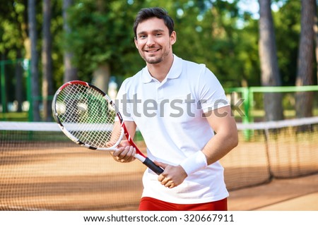 Picture of handsome young man on tennis court. Man playing tennis. Man cheerfully smiling, looking at camera and holding tennis racquet. Beautiful forest area as background