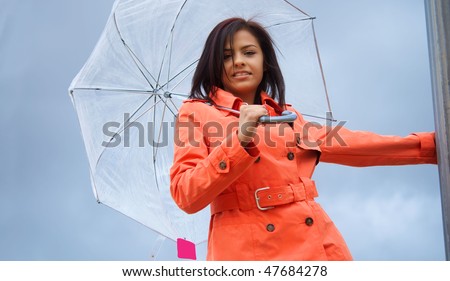 Beautiful young happy vibrant smiling young woman wearing an orange tench coat and umbrella on a cloudy rainy day.