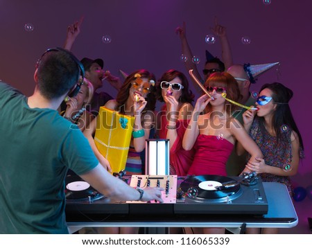 girls amusing themselves at party, dancing and playing, in front of dj mixing music, with poeple danging in the background