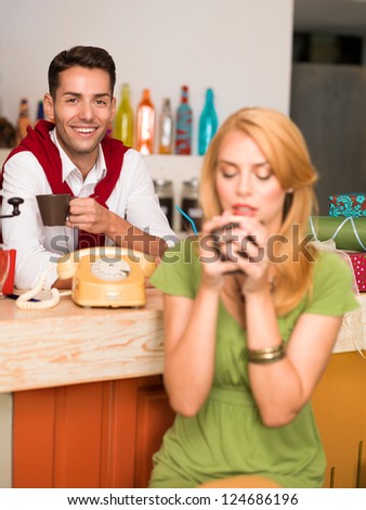 young handsome caucasian guy smiling with a cup of coffee in his hand, behind the counter of a colorful cafe, with a blonde girl enjoying her drink in front of him