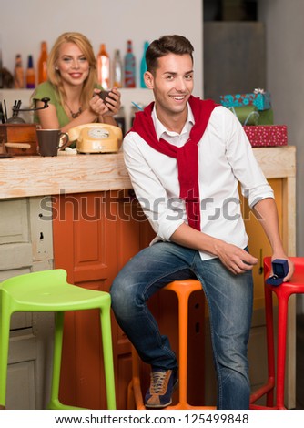 handsome caucasian man sitting on a chair at the counter of a cafe holding a box with an engagement ring in it, with a blonde girl behind the counter