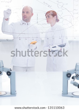 general-view  of a teacher in a chemistry lab drawing on a transparent board for his student in the background of a transparent board around lab table and lab tools