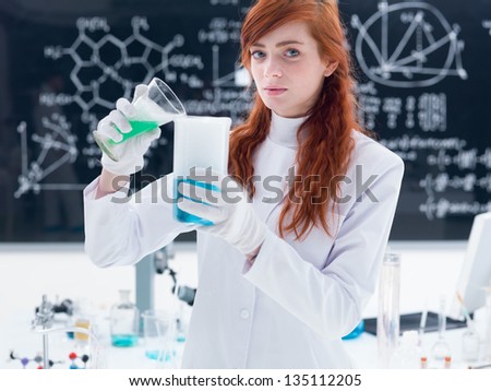 close-up of student in chemistry lab conducting an experiment with colorful liquids and magical gas around a lab table and looking in the camera  with a blackboard on the background