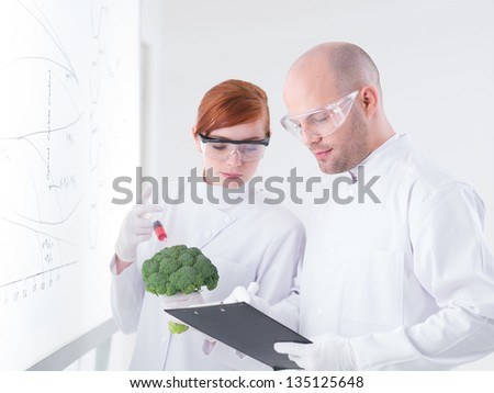 close-up of two people in a chemistry lab injecting a broccoli with a whiteboard on the background