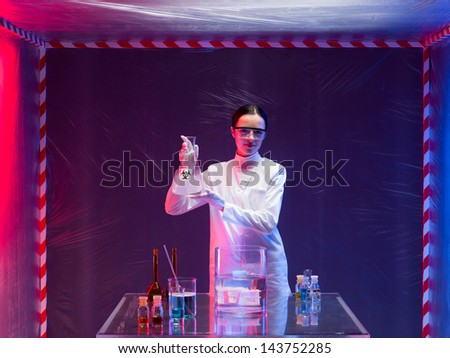 woman scientist wearing a white lab coat and protection goggles holding a bottle labeled bio hazardous with both hands in front of a table with glassware filled with chemicals, in a containment tent